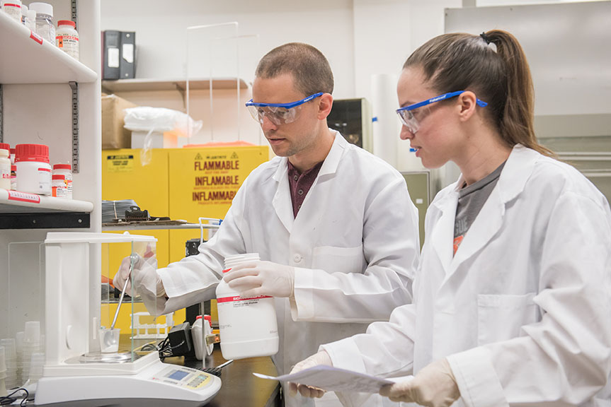 Two students working side-by-side in lab with chemicals and equipment