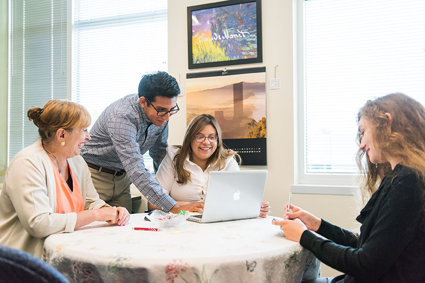 Four students gathered around a table while two work on a computer.