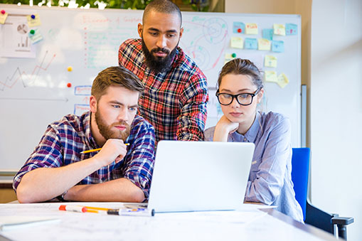 Three students focusing intensely on a computer screen with project notes in the background