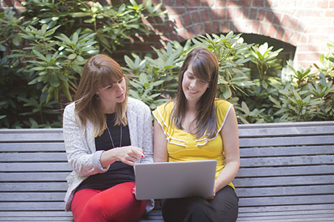 Two students sitting together on a bench working on a laptop.