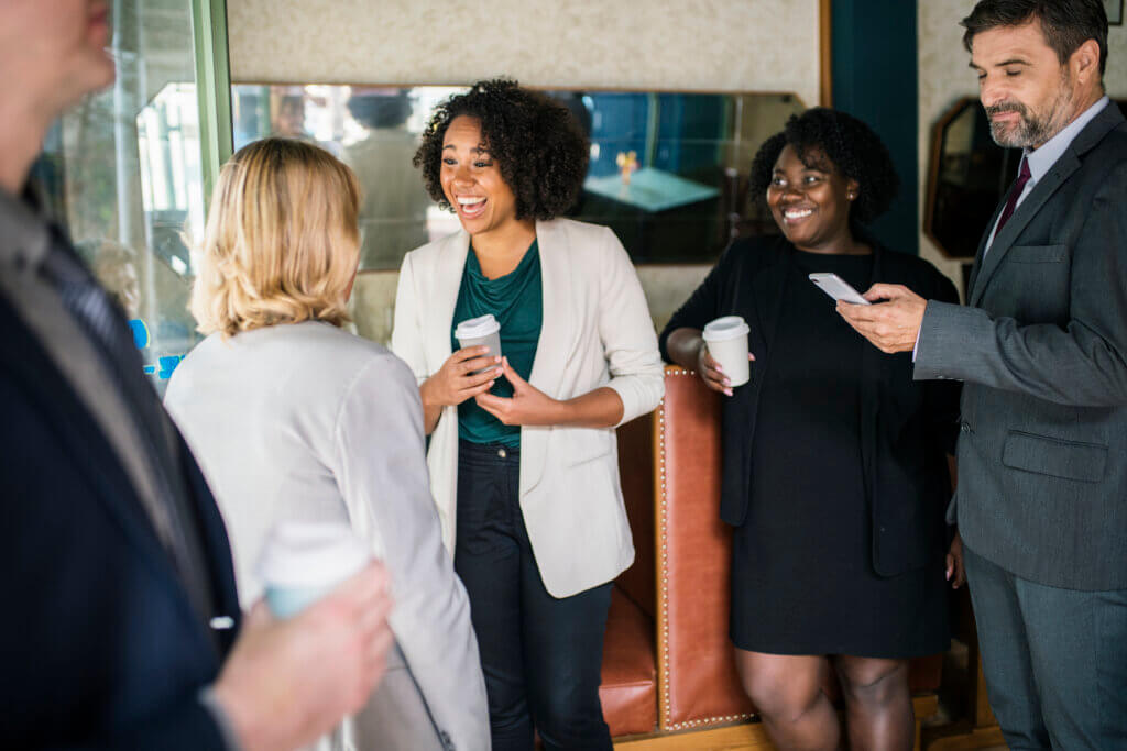 Three businesswomen smiling and having a lighthearted conversation in a modern office setting, possibly discussing an elevator pitch, highlighting a mix of professionalism and connection.