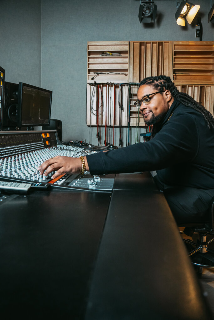 A man with long braids and glasses adjusts controls on a large audio mixing console in a professional recording studio.
