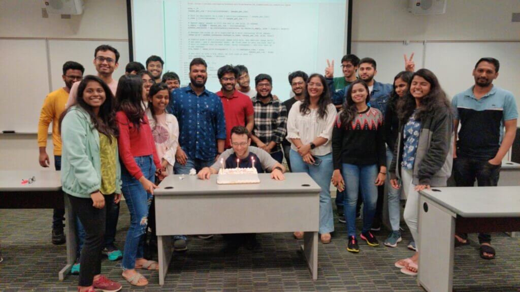 Group of UMBC Data Science students gathered around a classmate celebrating a birthday with a cake and candles in a classroom.