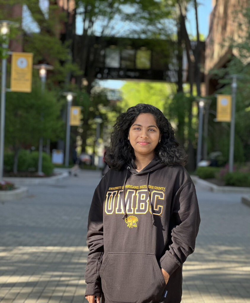 A young woman stands outdoors on a university campus wearing a black UMBC hoodie with hands in her pockets. She is looking at the camera with a slight smile. Trees and UMBC banners line the walkway behind her on a sunny day.
