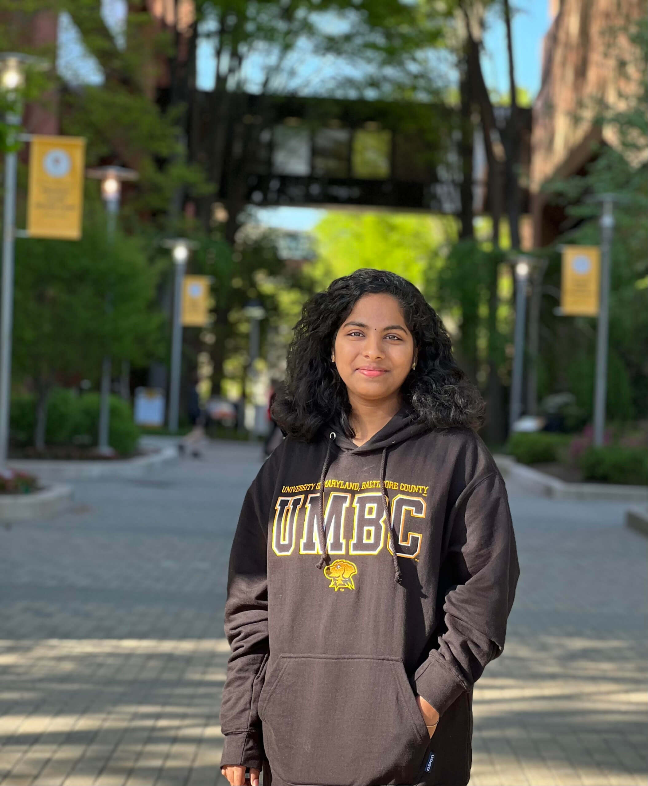 A young woman stands outdoors on a university campus wearing a black UMBC hoodie with hands in her pockets. She is looking at the camera with a slight smile. Trees and UMBC banners line the walkway behind her on a sunny day.