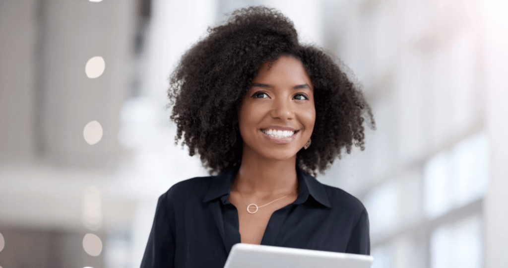 Smiling woman with curly hair wearing a dark blouse and holding a tablet in a bright indoor setting. The background is softly blurred, creating a professional and welcoming appearance.